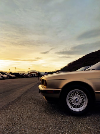 A classic car with a shiny gold exterior is parked on a paved road, with rows of other cars in the background. The sky is filled with a mixture of clouds and warm hues from the setting sun, casting a golden glow over the scene. Mountains can be seen in the distance, and a few people are standing nearby.