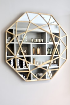 A geometric mirror with a gold frame reflects a set of shelves filled with decorative objects such as vases, canisters, and various containers. The shelves have a modern and minimalist design, displaying items in a symmetrical and organized manner. The reflected objects seem sophisticated and elegant.
