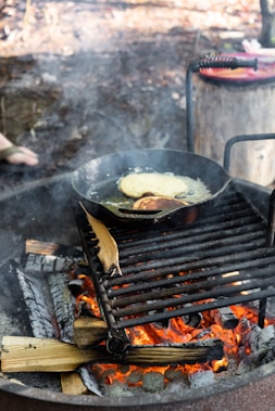 A cast-iron skillet sits on a metal grill over an open wood fire, cooking pancakes. The firewood underneath burns with bright orange flames, and there is visible smoke rising. A log or stump is seen in the background, possibly used as a seat or table, with a hint of a red object on top.