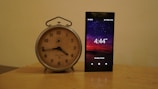 A vintage analog clock with a beige face and black hands is placed beside a modern smartphone displaying the time 4:44 PM against a backdrop of a starry night sky. The clock has a silver metal frame, while the phone screen shows detailed icons and the date Monday, 24 April.
