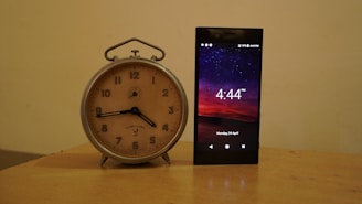 A vintage analog clock with a beige face and black hands is placed beside a modern smartphone displaying the time 4:44 PM against a backdrop of a starry night sky. The clock has a silver metal frame, while the phone screen shows detailed icons and the date Monday, 24 April.