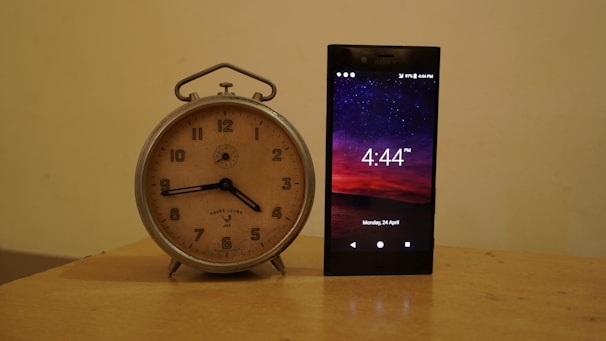 A vintage analog clock with a beige face and black hands is placed beside a modern smartphone displaying the time 4:44 PM against a backdrop of a starry night sky. The clock has a silver metal frame, while the phone screen shows detailed icons and the date Monday, 24 April.
