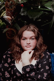 A person with curly hair and a nose ring, wearing a floral patterned top, is seated against a backdrop of lush green foliage and decorative elements.