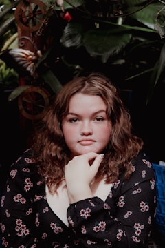 A person with curly hair and a nose ring, wearing a floral patterned top, is seated against a backdrop of lush green foliage and decorative elements.