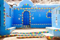 A vibrantly painted building with bright blue walls adorned with colorful geometric patterns in red, yellow, and green. It features a central blue door and colorful illustrations, including a crocodile and floral motifs. Arabic script and the word 'welcome' are visible, enhancing the cultural feel of the setting.