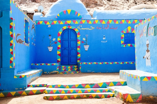 A vibrantly painted building with bright blue walls adorned with colorful geometric patterns in red, yellow, and green. It features a central blue door and colorful illustrations, including a crocodile and floral motifs. Arabic script and the word 'welcome' are visible, enhancing the cultural feel of the setting.