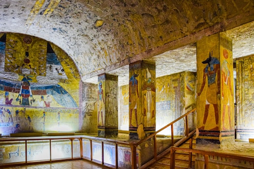 An ancient Egyptian tomb with vivid wall paintings and hieroglyphics. The space includes decorated columns featuring Egyptian deities and intricate artwork. The lighting highlights the vibrant colors on the walls, enhancing the detail of the historical art.