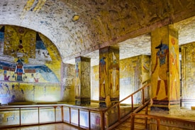 An ancient Egyptian tomb with vivid wall paintings and hieroglyphics. The space includes decorated columns featuring Egyptian deities and intricate artwork. The lighting highlights the vibrant colors on the walls, enhancing the detail of the historical art.