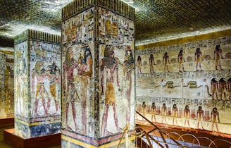 The walls and columns are adorned with intricate ancient Egyptian hieroglyphs and colorful paintings. Deities and figures are depicted in traditional Egyptian attire, showcasing scenes that appear to tell a story or represent significant cultural or religious events. The ceiling is decorated with a pattern resembling the night sky.
