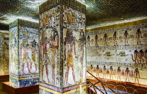 The walls and columns are adorned with intricate ancient Egyptian hieroglyphs and colorful paintings. Deities and figures are depicted in traditional Egyptian attire, showcasing scenes that appear to tell a story or represent significant cultural or religious events. The ceiling is decorated with a pattern resembling the night sky.