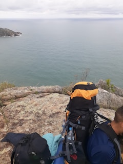 A scenic view of an ocean with a rocky outcrop in the foreground. There is a person wearing a backpack with camping gear, including a sleeping bag and water bottle, sitting to the side. The sky is overcast, creating a calm and serene atmosphere.