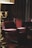 A dimly lit, cozy room featuring a large, plush leather armchair in the foreground. The armchair is a deep red color, complementing the wooden panels and dark-toned furniture surrounding it. A lamp with a gold-toned base adds a warm glow to the scene. Several high-backed chairs are visible around a polished wooden table in the background.