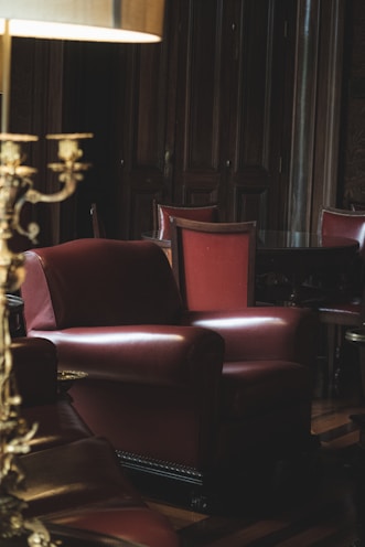 A dimly lit, cozy room featuring a large, plush leather armchair in the foreground. The armchair is a deep red color, complementing the wooden panels and dark-toned furniture surrounding it. A lamp with a gold-toned base adds a warm glow to the scene. Several high-backed chairs are visible around a polished wooden table in the background.