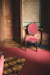 A richly decorated room with a single upholstered chair in red fabric and wooden frame. The walls feature ornate wood paneling and a patterned wallpaper in muted colors. A carpet in the same red shade as the chair covers the floor, while an intricate wooden inlay adds detail to the remaining floor space.