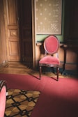 A richly decorated room with a single upholstered chair in red fabric and wooden frame. The walls feature ornate wood paneling and a patterned wallpaper in muted colors. A carpet in the same red shade as the chair covers the floor, while an intricate wooden inlay adds detail to the remaining floor space.