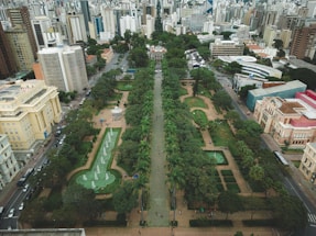 Aerial view of a large urban park surrounded by numerous tall buildings in a city. The park features symmetrical landscaping with green lawns, pathways, and several fountains. Trees line the central pathway, which is flanked by lush greenery, small gardens, and pedestrian areas. The surrounding area consists of a mix of modern and historic buildings with a variety of architectural styles.