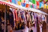 Mannequins displaying a variety of colorful, patterned garments under a canopy with vibrant paper decorations hanging above. The setting appears to be an outdoor market with clothes in traditional or festive designs.
