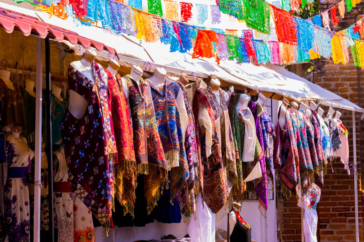 Mannequins displaying a variety of colorful, patterned garments under a canopy with vibrant paper decorations hanging above. The setting appears to be an outdoor market with clothes in traditional or festive designs.