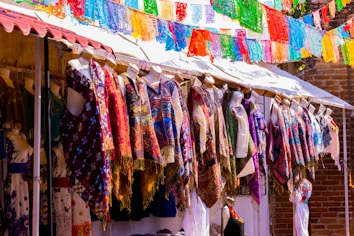 Mannequins displaying a variety of colorful, patterned garments under a canopy with vibrant paper decorations hanging above. The setting appears to be an outdoor market with clothes in traditional or festive designs.