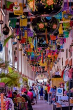 A vibrant market scene filled with colorful decorations hanging from the ceiling, including papel picado and painted gourds. Numerous stalls line the corridor, displaying a variety of handmade crafts and textiles. Many people, including vendors and shoppers, fill the bustling space.
