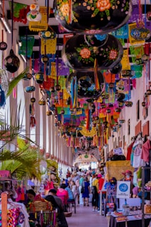 A vibrant market scene filled with colorful decorations hanging from the ceiling, including papel picado and painted gourds. Numerous stalls line the corridor, displaying a variety of handmade crafts and textiles. Many people, including vendors and shoppers, fill the bustling space.