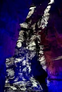 Black and white image of soldiers appears projected onto rough, rocky cave walls, creating a historical and ghostly effect. The distinct contrast between the monochrome figures and the vibrant blue and purple lighting surrounding them creates a surreal, immersive environment.