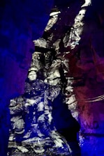 Black and white image of soldiers appears projected onto rough, rocky cave walls, creating a historical and ghostly effect. The distinct contrast between the monochrome figures and the vibrant blue and purple lighting surrounding them creates a surreal, immersive environment.