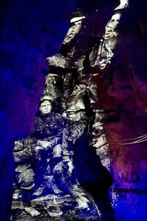 Black and white image of soldiers appears projected onto rough, rocky cave walls, creating a historical and ghostly effect. The distinct contrast between the monochrome figures and the vibrant blue and purple lighting surrounding them creates a surreal, immersive environment.