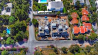 An aerial view of a residential area shows a construction site in the center, surrounded by roads and several houses with red roofs. The site features partially built structures with exposed concrete and construction materials. Greenery and trees fill the surrounding landscape, along with additional houses featuring swimming pools.