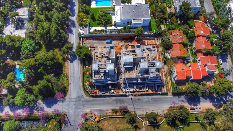An aerial view of a residential area shows a construction site in the center, surrounded by roads and several houses with red roofs. The site features partially built structures with exposed concrete and construction materials. Greenery and trees fill the surrounding landscape, along with additional houses featuring swimming pools.