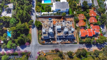 An aerial view of a residential area shows a construction site in the center, surrounded by roads and several houses with red roofs. The site features partially built structures with exposed concrete and construction materials. Greenery and trees fill the surrounding landscape, along with additional houses featuring swimming pools.