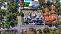 An aerial view of a residential area shows a construction site in the center, surrounded by roads and several houses with red roofs. The site features partially built structures with exposed concrete and construction materials. Greenery and trees fill the surrounding landscape, along with additional houses featuring swimming pools.