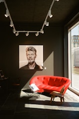 A modern interior with a striking black wall features a portrait of a person and a vivid red couch. The room is illuminated by track lighting and a large window allows natural light to stream in. A small table and another chair are also visible, adding to the minimalist yet stylish decor.