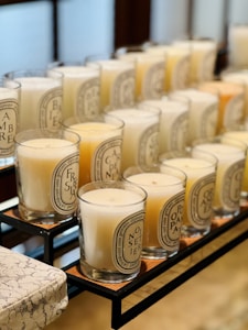 A neatly arranged collection of scented candles in glass jars, each featuring distinct labels with intricate lettering. The candles are placed on a metal rack, with varied wax colors ranging from white to light yellow and pale orange. The setting is indoors, with a blurred background providing a warm ambiance.
