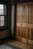 A wooden wardrobe with paneled doors is adjacent to a window with dark wooden frames. The interior is warmly lit, highlighting the natural grain of the wood. A patterned rug lies on the wooden floor.