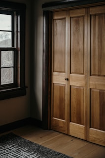 A wooden wardrobe with paneled doors is adjacent to a window with dark wooden frames. The interior is warmly lit, highlighting the natural grain of the wood. A patterned rug lies on the wooden floor.