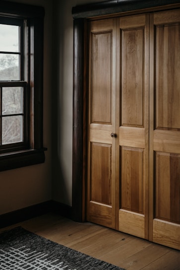 A wooden wardrobe with paneled doors is adjacent to a window with dark wooden frames. The interior is warmly lit, highlighting the natural grain of the wood. A patterned rug lies on the wooden floor.