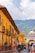 A colorful street scene in a historic town with vibrant yellow and orange buildings lining the street. The image captures a lively atmosphere with people walking and a motorbike passing by. The architecture showcases traditional design with decorative lamps and wooden elements. In the background, lush green mountains provide a scenic backdrop under a partly cloudy sky.