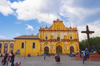 A large, ornate yellow and red colonial-style cathedral stands prominently in an open plaza under a partly cloudy blue sky. People walk and gather around, with a vendor selling colorful balloons on the left. A large wooden cross is located on the right side of the image, with several people sitting around its base.