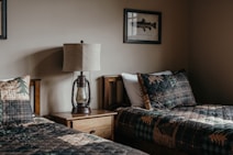 A cozy bedroom scene features two twin beds with plaid patterned bedding. A wooden nightstand is positioned between the beds, topped with a rustic lamp resembling an oil lantern. On the walls are framed art pieces, one depicting a fish. The overall ambiance is warm and inviting.