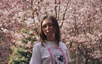A woman with straight brown hair stands in front of a backdrop of blooming cherry blossoms. She is wearing a white t-shirt featuring an anime character. The sunlight softly illuminates her face as she seems lost in thought with her eyes closed or looking down.