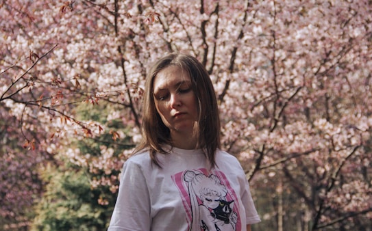 A woman with straight brown hair stands in front of a backdrop of blooming cherry blossoms. She is wearing a white t-shirt featuring an anime character. The sunlight softly illuminates her face as she seems lost in thought with her eyes closed or looking down.