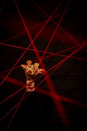 A plush giraffe toy is illuminated against a dark background with several intersecting red laser beams. The giraffe is positioned upright, appearing playful amidst the stark contrast of the lasers.