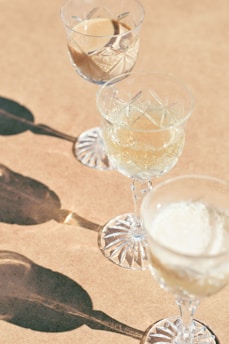 Three crystal glasses filled with different liquids are placed on a light brown surface. The glasses cast distinct, elongated shadows, adding an artistic flair to the composition. The glassware features intricate cut patterns, suggesting elegance and sophistication.