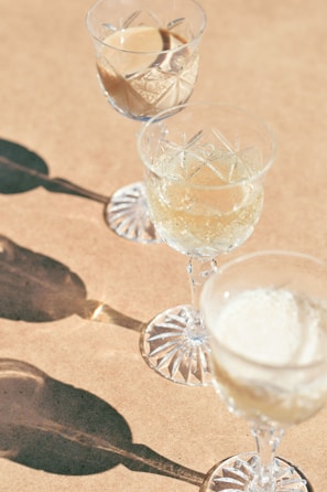 Three crystal glasses filled with different liquids are placed on a light brown surface. The glasses cast distinct, elongated shadows, adding an artistic flair to the composition. The glassware features intricate cut patterns, suggesting elegance and sophistication.