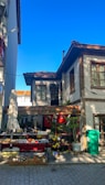 A small outdoor market stall is set up in front of a rustic two-story house with a tiled roof and wooden accents. Various colorful trinkets and goods are displayed on tables. A large green recycling bin is situated to the right. The bright blue sky is visible above.