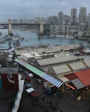 A bustling public market complex located on Granville Island near a waterfront with boats and a bridge in the background. The market buildings are topped with colorful awnings, and high-rise buildings dominate the skyline. The scene appears overcast and wet, suggesting a rainy day.