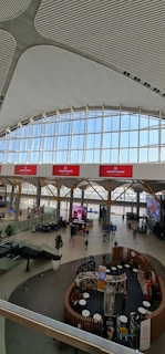 The image displays the interior of a large, modern airport terminal with a high, patterned ceiling and expansive windows allowing natural light in. There are red digital displays with airline branding, and passengers are scattered throughout the spacious terminal. Various seating arrangements and informational kiosks are present, along with a few green potted plants for decoration.