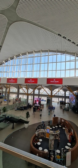 The image displays the interior of a large, modern airport terminal with a high, patterned ceiling and expansive windows allowing natural light in. There are red digital displays with airline branding, and passengers are scattered throughout the spacious terminal. Various seating arrangements and informational kiosks are present, along with a few green potted plants for decoration.