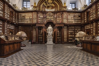 A grand, classical library interior featuring ornate wooden bookshelves filled with books that line the walls. At the center stands a prominent statue of a historical figure surrounded by architectural details like columns and arches. Two large, antique globes are positioned on either side of the room, and a checkered floor adds to the elegant ambiance.